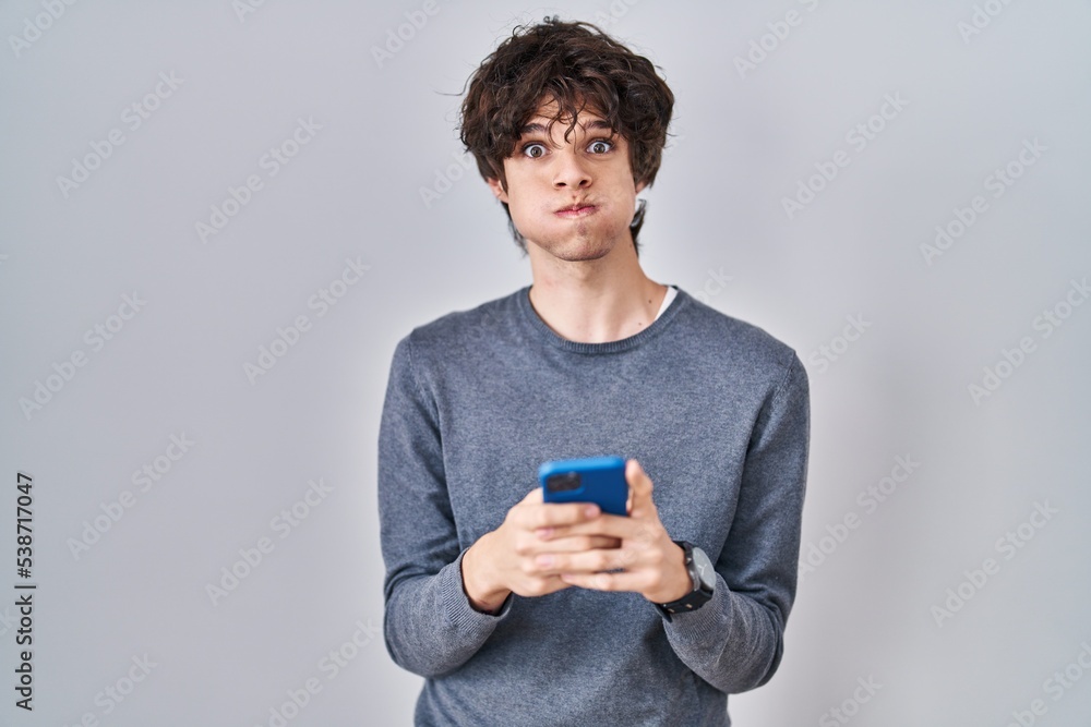 Young man using smartphone typing a message puffing cheeks with funny face. mouth inflated with air, catching air.