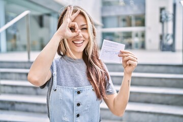  Young caucasian woman holding covid record card smiling happy doing ok sign with hand on eye looking through fingers