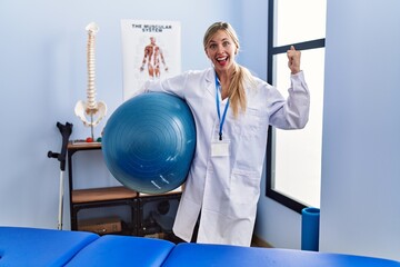 Poster - Beautiful woman holding pilates ball at physiotherapy clinic screaming proud, celebrating victory and success very excited with raised arm