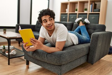 Poster - Young hispanic man reading book lying on the sofa at home.