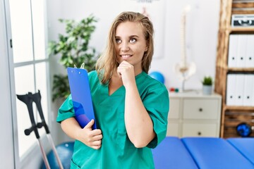 Poster - Young caucasian woman working at pain recovery clinic with hand on chin thinking about question, pensive expression. smiling and thoughtful face. doubt concept.