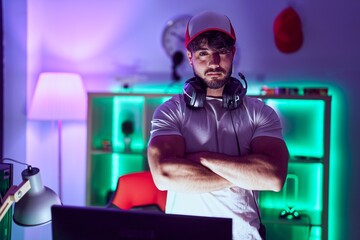 Young hispanic man streamer standing with arms crossed gesture at gamin room