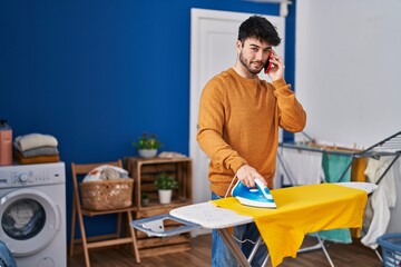 Poster - Young hispanic man talking on the smartphone ironing clothes at laundry room