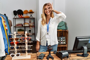 Canvas Print - Blonde beautiful young woman working as manager at retail boutique doing ok gesture with hand smiling, eye looking through fingers with happy face.