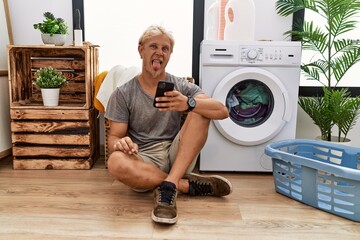 Poster - Young blond man doing laundry using smartphone sticking tongue out happy with funny expression. emotion concept.