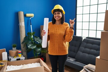 Wall Mural - Young hispanic woman standing on new home with blueprints smiling happy pointing with hand and finger to the side