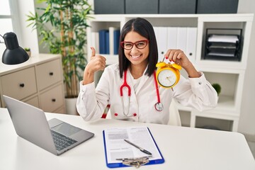 Canvas Print - Young hispanic doctor woman holding alarm clock at the clinic smiling happy and positive, thumb up doing excellent and approval sign