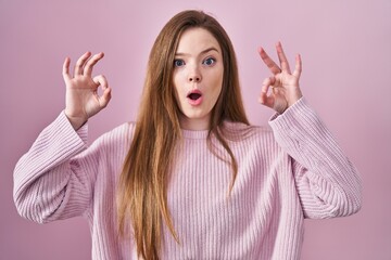 Poster - Young caucasian woman standing over pink background looking surprised and shocked doing ok approval symbol with fingers. crazy expression