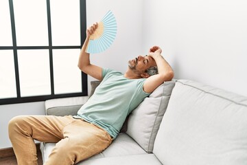 Wall Mural - Middle age grey-haired man using handfan sitting on sofa at home