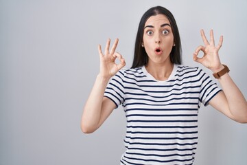 Wall Mural - Young brunette woman wearing striped t shirt looking surprised and shocked doing ok approval symbol with fingers. crazy expression