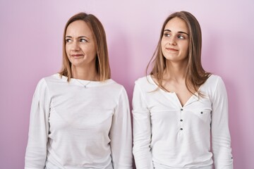 Wall Mural - Middle age mother and young daughter standing over pink background smiling looking to the side and staring away thinking.