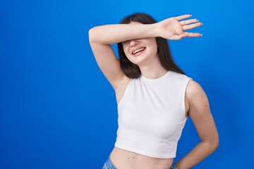 Canvas Print - Young caucasian woman standing over blue background covering eyes with arm smiling cheerful and funny. blind concept.