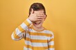 Young caucasian kid standing over yellow background smiling and laughing with hand on face covering eyes for surprise. blind concept.