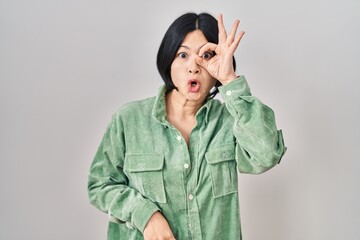 Poster - Young asian woman standing over white background doing ok gesture shocked with surprised face, eye looking through fingers. unbelieving expression.