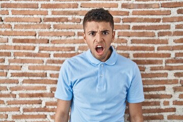 Sticker - Brazilian young man standing over brick wall in shock face, looking skeptical and sarcastic, surprised with open mouth