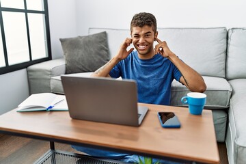 Poster - Young handsome hispanic man using laptop sitting on the floor covering ears with fingers with annoyed expression for the noise of loud music. deaf concept.