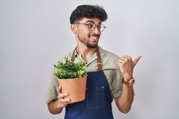 Poster - Arab man with beard holding green plant pot smiling with happy face looking and pointing to the side with thumb up.