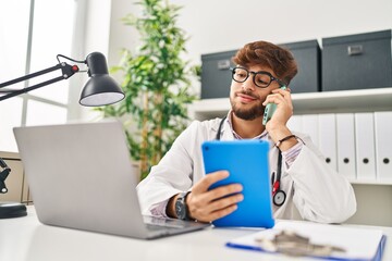 Canvas Print - Young arab man wearing doctor uniform talking on smartphone using touchpad at clinic