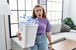 Young redhead woman holding recycling wastebasket with plastic bottles in shock face, looking skeptical and sarcastic, surprised with open mouth