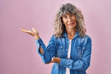 Canvas Print - Middle age woman standing over pink background smiling cheerful presenting and pointing with palm of hand looking at the camera.