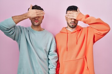 Wall Mural - Young hispanic gay couple standing over pink background covering eyes with hand, looking serious and sad. sightless, hiding and rejection concept