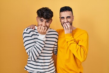 Poster - Young hispanic gay couple standing over yellow background laughing and embarrassed giggle covering mouth with hands, gossip and scandal concept