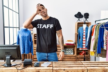 Wall Mural - Young hispanic man working at retail boutique doing ok gesture shocked with surprised face, eye looking through fingers. unbelieving expression.