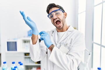 Poster - Young hispanic man working at scientist laboratory putting gloves on celebrating crazy and amazed for success with open eyes screaming excited.