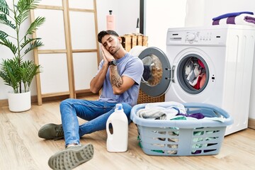 Wall Mural - Young hispanic man putting dirty laundry into washing machine sleeping tired dreaming and posing with hands together while smiling with closed eyes.