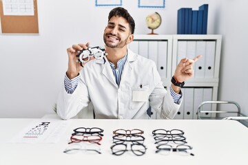 Wall Mural - Young optician man holding optometry glasses cheerful with a smile on face pointing with hand and finger up to the side with happy and natural expression