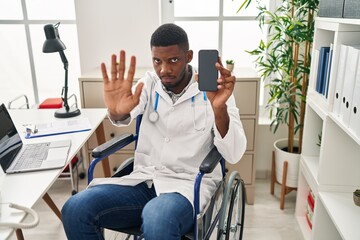 Poster - African american doctor man sitting on wheelchair holding smartphone with open hand doing stop sign with serious and confident expression, defense gesture