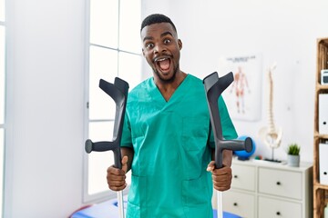 Wall Mural - Young african physiotherapist man holding crutches celebrating crazy and amazed for success with open eyes screaming excited.