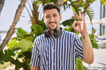 Canvas Print - Young handsome man listening to music using headphones outdoors smiling positive doing ok sign with hand and fingers. successful expression.