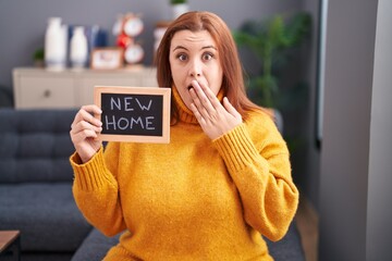 Young hispanic woman with red hair holding blackboard with new home text covering mouth with hand, shocked and afraid for mistake. surprised expression