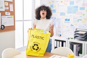 Canvas Print - Young middle east woman holding recycling bag with plastic bottles at the office angry and mad screaming frustrated and furious, shouting with anger looking up.