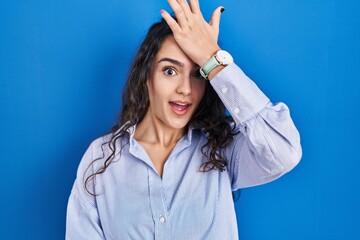 Poster - Young brunette woman standing over blue background surprised with hand on head for mistake, remember error. forgot, bad memory concept.