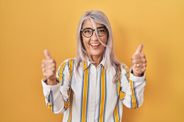 Wall Mural - Middle age woman with grey hair standing over yellow background wearing glasses success sign doing positive gesture with hand, thumbs up smiling and happy. cheerful expression and winner gesture.