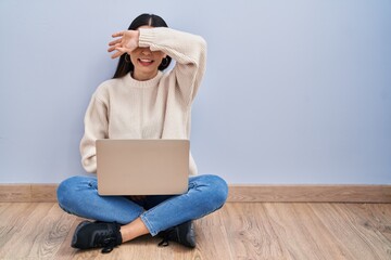 Sticker - Young woman using laptop sitting on the floor at home covering eyes with arm smiling cheerful and funny. blind concept.