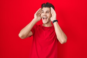 Poster - Young hispanic man standing over red background smiling cheerful playing peek a boo with hands showing face. surprised and exited