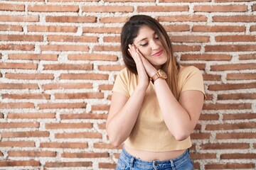 Wall Mural - Young brunette woman standing over bricks wall sleeping tired dreaming and posing with hands together while smiling with closed eyes.