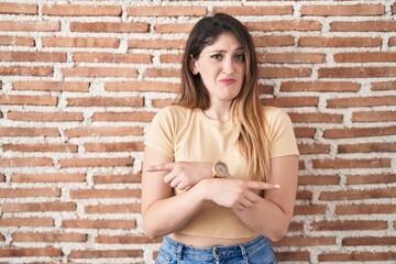 Poster - Young brunette woman standing over bricks wall pointing to both sides with fingers, different direction disagree