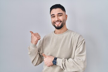 Sticker - Young handsome man standing over isolated background pointing to the back behind with hand and thumbs up, smiling confident