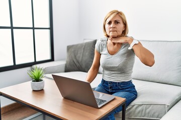 Poster - Middle age blonde woman using laptop at home cutting throat with hand as knife, threaten aggression with furious violence