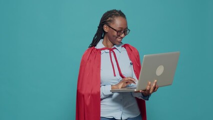 Woman dressed as superhero using laptop in studio, wearing red cape hero costume and browsing internet. Looking at computer with online network, having cloak and cartoon comic look.