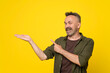Middle-age handsome man wearing casual green shirt standing over isolated yellow background smiling cheerful pointing with hand and finger up to the side to open horizontal palm gesture