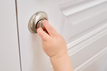 Wall Mural - Toddler baby opens the lock, child hand close-up. White wooden door, metal door handle and baby hand