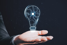Close Up Of Businessman Hand Holding Abstract Glowing Light Bulb With Chip Hologram On Blurry Dark Background. Artificial Intelligence And Neural Network Concept.