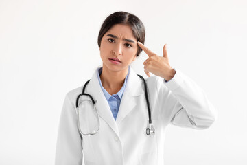 Wall Mural - Tired sad millennial indian woman doctor in coat show hand gesture, pistol near temple