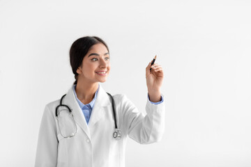 Wall Mural - Cheerful millennial hindu lady therapist in coat writes with pen on empty space