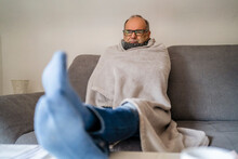 Senior Man Wrapped In Blanket Sitting On Sofa At Home